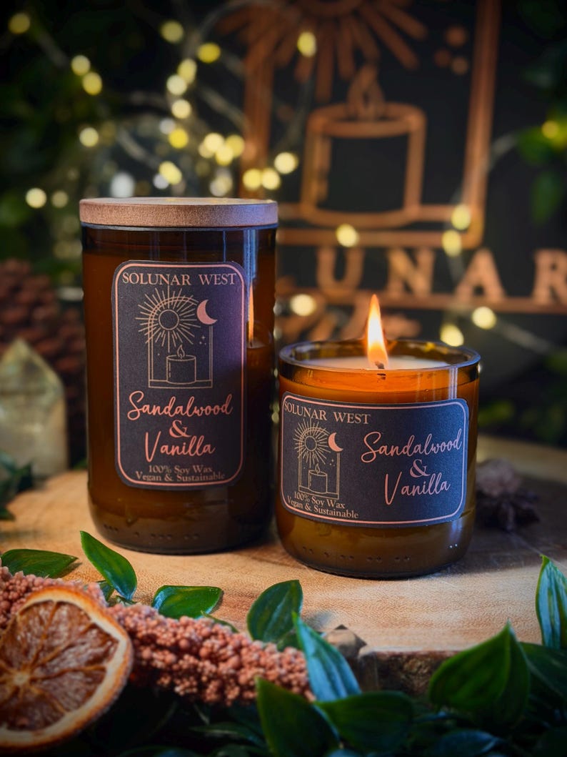 May include: Two glass jar candles with wooden lids, labeled "Sandalwood & Vanilla" by Solunar West. The candles are on a wooden surface with dried orange slices and greenery. One candle is lit, showing a flame.