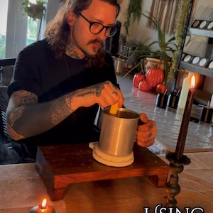 May include: A metal pouring pitcher for candle making, a skull-shaped candle, and a lit taper candle. Text reads: ALL CANDLES ARE HANDMADE IN WEST WALES USING 100% SOY WAX & TOXIC FREE SCENTS. The scene includes a wooden stand and a burning incense stick.