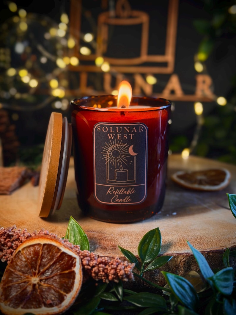 May include: A lit candle in a dark amber glass jar with a wooden lid. The label features the text "SOLUNAR WEST" and "Refillable Candle" with a graphic of a sun, moon, and candle. The flame is visible, and the jar has a deep, warm tone.