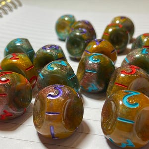 Puede incluir: Una colección de dados moteados multicolores. Los dados son principalmente tonos de marrón, verde y dorado, con números en rojo, azul y morado. Los dados están dispuestos sobre una superficie blanca.