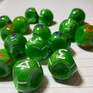May include: A group of 12 green dice with white numbers and orange and purple accents. The dice are scattered on a white surface with lined paper.