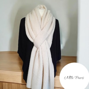 May include: A cream-colored, chunky knit scarf draped over a black top. The scarf is thick and textured, with a ribbed pattern. The scarf is styled in a crossed, wrapped design. The background is a neutral color. The brand name "Mlle Paris" is in the lower right corner.