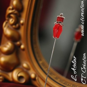 May include: A red hat pin with a silver stem, positioned in front of a gold-framed mirror. The pin features a faceted red gem and a small hat design. The mirror reflects the pin and the text "www.ctcreation.fr".