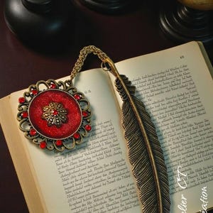 Könnte beinhalten: Dekorativer Lesezeichen mit rotem und goldenem Medaillon und Federdesign. Das Medaillon hat rote Edelsteine und ein Blumenmuster. Das Lesezeichen liegt auf einem offenen Buch.