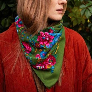 May include: A green scarf with a floral pattern featuring pink, blue, and yellow flowers. The scarf is worn around the neck and is set against a red jacket. The scarf's design is a vibrant contrast to the solid-colored jacket.