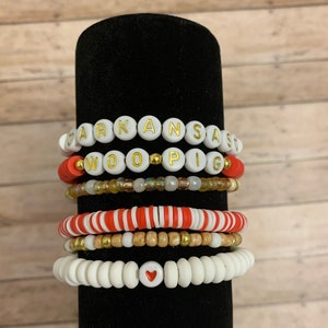 May include: Stack of six beaded bracelets in various designs. The top bracelet spells "ARKANSAS" in gold letters on white beads. Other bracelets feature red, white, and gold beads, with one displaying a heart symbol.