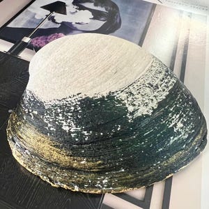 May include: A large, decorative seashell painted with a gradient of colors. The top half is white, transitioning to dark green and gold at the base. The shell is a unique piece of home decor.