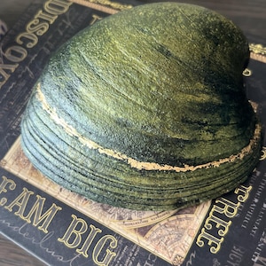 May include: A large, decorative clam shell with a textured, green and gold finish. The shell is resting on a book with the words "DREAM BIG" and "DISCOVER" in gold lettering. The shell's ridges are highlighted with gold.