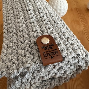 Op de afbeelding: Een gevouwen, lichtgrijze gehaakte deken met een gestructureerd steekpatroon. Een bruin leren label met de woorden "HANDMADE WITH LOVE BY WOLLE UND LIEBE" is bevestigd. Een witte vaas staat op de achtergrond.