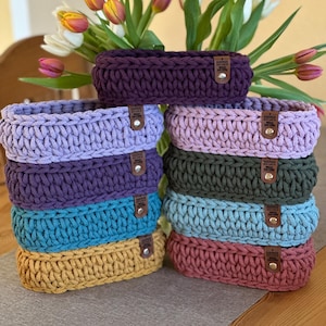 Könnte beinhalten: Ein Stapel von acht gehäkelten Aufbewahrungskörben in verschiedenen Farben, darunter Lila, Lavendel, Türkis, Senfgelb und Rot. Jeder Korb hat ein braunes Leder-Etikett mit dem Wort "Handmade" darauf.
