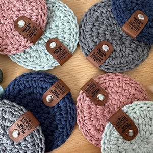 Könnte beinhalten: Acht gehäkelte Untersetzer in verschiedenen Farben, darunter Rosa, Grau, Blau und Grün. Jeder Untersetzer hat ein braunes Leder-Etikett mit dem Text "HANDMADE WITH LOVE BY WOLLE UND LIEBE".