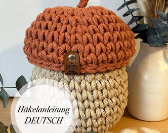 May include: A crocheted acorn with a brown top and a beige bottom. The acorn has a small stem on top. The text "Handmade with love" is on a brown leather tag attached to the acorn. The text "Häckelanleitung DEUTSCH" is in a white circle on a green leafy background.