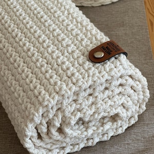 Könnte beinhalten: Eine aufgerollte, cremefarbene Häkeldecke, die mit einem braunen Lederriemen befestigt ist. Der Riemen ist mit einem silbernen Druckknopf befestigt und hat einen geprägten Text. Ein Teil eines passenden Häkelkorbs ist sichtbar.