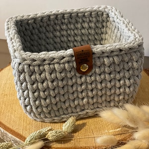 Peut inclure: Un panier de rangement en crochet gris avec une étiquette en cuir marron qui dit "Handmade with love by" et un bouton doré. Le panier est posé sur une surface en bois.