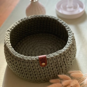 Può includere: Un cesto in crochet verde salvia con un'etichetta in pelle marrone che dice "Handmade with love".