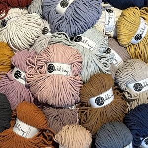 Könnte beinhalten: Eine Auswahl an Bobbiny Makramee-Garn in verschiedenen Farben wie Creme, Rosa, Braun und Blau. Jede Garnrolle ist eng gewickelt und mit dem Markennamen versehen. Geeignet für Bastelprojekte.