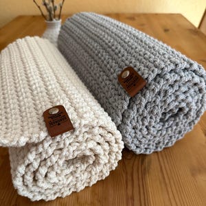 Puede incluir: Dos mantas de punto grueso enrolladas. Una es blanquecina, la otra gris. Cada manta tiene una etiqueta de cuero marrón con la palabra "HANDMADE". Las mantas están sobre una superficie de madera.