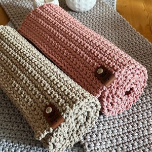 Peut inclure: Deux chemins de table en crochet roulés, beige et rose. Les chemins de table sont posés sur un chemin de table en crochet gris. Chaque chemin de table roulé a une petite étiquette en cuir marron. Un vase blanc et une bougie sont en arrière-plan.