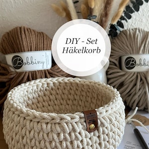 Könnte beinhalten: Ein runder, cremefarbener Häkelkorb aus dickem Garn mit einem kleinen braunen Lederetikett mit der Aufschrift 'Handmade with love by Wolle und Liebe'. Dahinter liegen zwei Bobbiny-Garnknäuel und ein gedrucktes Anleitungsblatt.