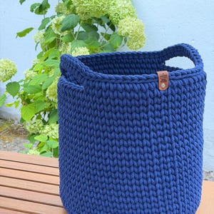 May include: A deep navy blue cylindrical crochet basket featuring a thick, textured knit pattern and two integrated handles. A small, rectangular brown leather tag is attached to the upper rim, embossed with the text "HANDMADE" and "MADE WITH LOVE".