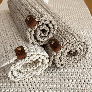 Könnte beinhalten: Drei gerollte, handgefertigte, beige gehäkelte Tischsets mit braunen Lederetiketten mit der Aufschrift "HANDMADE". Die Tischsets liegen auf einer größeren, passenden Häkeloberfläche. Die Textur ist sichtbar, und die Farbpalette ist neutral.