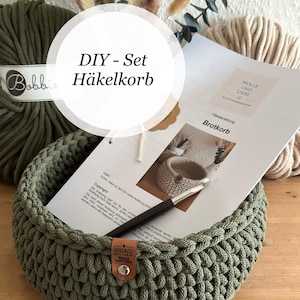Könnte beinhalten: Ein DIY-Häkelset bestehend aus einem runden, olivgrünen Häkelkorb mit braunem Lederetikett, einer gedruckten Anleitung mit dem Titel Brotkorb, einer Metallhäkelnadel und einem großen Knäuel olivgrünem Garn im Hintergrund.