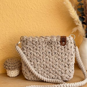 Op de afbeelding: Een beige gehaakte schoudertas met een stervormig steekpatroon, een lange bijpassende riem en een klein bruin leren label in de rechterbovenhoek met de tekst "HANDMADE WITH LOVE BY WOLLE UND GARN".