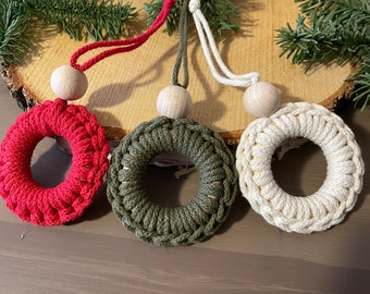 May include: Three macrame wreath ornaments in red, olive green, and cream colors. Each wreath has a wooden bead at the top and a matching cord for hanging. The wreaths are displayed on a wooden slice with evergreen sprigs.