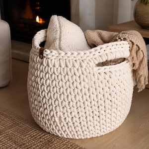 May include: A round, off-white chunky knit storage basket made of thick cord, featuring integrated side handles and a small brown leather accent tab. The basket contains a textured cream-coloured pillow and a beige fringed throw blanket.