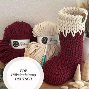 Könnte beinhalten: Ein handgefertigter Weihnachtsstrumpf in Burgunderrot und Creme, mit passendem Garn und einem Häkelhaken. Der Strumpf ist aus dickem, strukturiertem Garn gefertigt. Das Bild enthält auch zwei Knäuel Garn und den Text "PDF Häkelanleitung DEUTSCH".