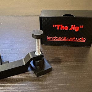 May include: A black 3D printed jig with a screw and a knob, labeled "The Jig" and "kindsaltystudio" in red text on a black box.