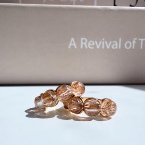 May include: A ring featuring a series of round, translucent, light brown rutilated quartz beads set in a gold-toned metal band. The beads display visible needle-like inclusions within the stone, creating a textured, shimmering appearance.