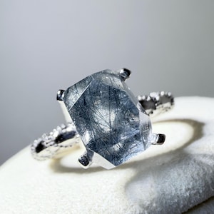 May include: A silver ring featuring a large, rectangular, faceted rutilated quartz gemstone with visible dark, needle-like inclusions throughout the stone. The metal band has a textured, uneven surface and is held by four prongs.