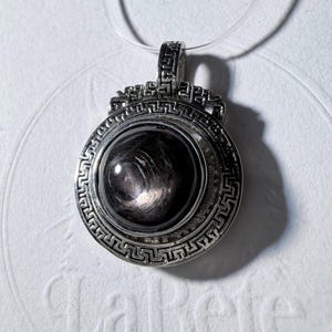 May include: A circular silver-toned pendant featuring a polished, dark, swirling gemstone at its center. The metal frame is decorated with a detailed Greek key pattern around the perimeter and a matching geometric design on the top bail.