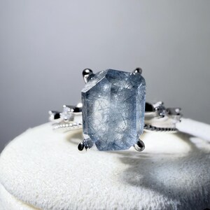 May include: A silver ring featuring a large, rectangular, faceted blue rutilated quartz gemstone with visible dark needle-like inclusions. The band is thin and metallic, accented with small, clear, round stones on either side of the center stone.