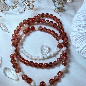 Puede incluir: Tres pulseras de cuentas con una mezcla de cuentas marrón rojizo y blancas. Las pulseras están dispuestas en capas, con un cristal transparente y flores secas en el fondo. Las cuentas son redondas y parecen ser de piedra.