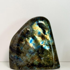 May include: A polished labradorite stone with a rounded top and a flat base. The stone displays iridescent flashes of blue, gold, and green within a dark gray matrix. The overall shape is smooth and organic.