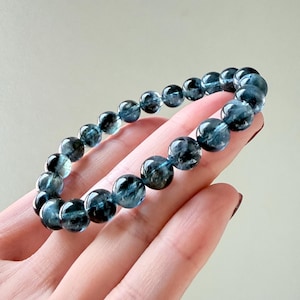 May include: A stretch bracelet composed of round, polished beads in varying shades of deep blue and dark teal. The beads exhibit a translucent quality with internal inclusions and dark, cloudy patterns throughout the spherical stone surfaces.