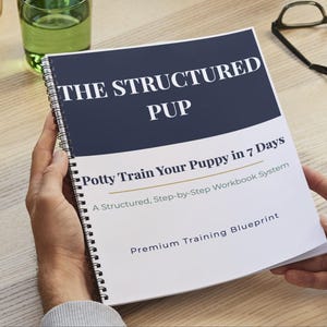 May include: A spiral-bound workbook titled "THE STRUCTURED PUP" with the subtitle "Potty Train Your Puppy in 7 Days." The cover is white with a navy blue header. The book is held open by a person's hands on a light wood surface.