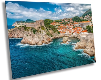May include: A scenic view of a coastal city with a fortress on a rocky peninsula. The turquoise water contrasts with the orange rooftops and stone walls. The sky is blue with fluffy white clouds.