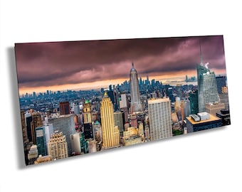 May include: A panoramic view of the New York City skyline featuring the Empire State Building and various skyscrapers under a dramatic, dark, and cloudy sunset sky with orange light glowing on the horizon behind the dense urban architecture.