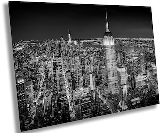 May include: A black and white wall art print featuring an aerial night view of the New York City skyline. The Empire State Building stands prominently in the foreground, surrounded by a dense grid of illuminated skyscrapers and city streets.