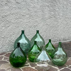 May include: A collection of large, glass demijohns in various shades of green and clear. The bottles have a rounded body and a narrow neck. They are arranged on a stone surface against a textured wall.