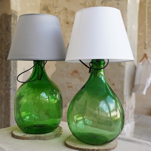 Peut inclure: Deux bouteilles en verre vert avec des abat-jours blancs sur le dessus. Les bouteilles sont sur des bases en bois et ont un aspect rustique.