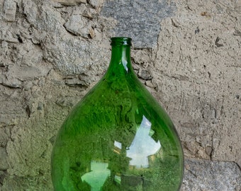 Puede incluir: Una botella de vidrio grande, redonda y verde con cuello estrecho. La botella está vacía y tiene un ligero reflejo del cielo.