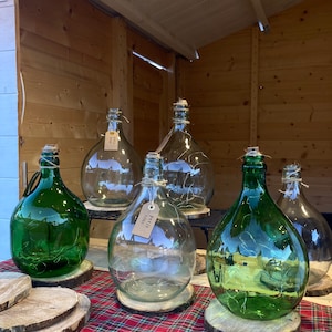 Peut inclure: Cinq bouteilles en verre avec des lumières féériques à l'intérieur, disposées sur des rondins de bois. Les bouteilles sont transparentes et vertes, certaines portant des étiquettes en papier brun avec du texte. Les bouteilles sont sur une nappe à carreaux rouge et noir.