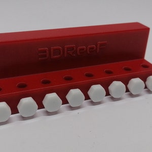 May include: A red plastic holder with the text "3DReef" printed on it. The holder has ten holes and white plastic pegs inserted into the holes.