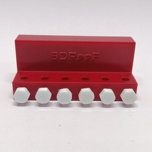 Op de afbeelding: Een rode plastic organizer met de tekst "3D FORCE" gedrukt op de voorkant. De organizer heeft acht gaten en houdt acht witte plastic zeskantmoeren vast.