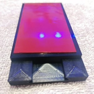 May include: A black 3D printed phone stand with a red adhesive pad on top. The stand has two triangular supports on the bottom.