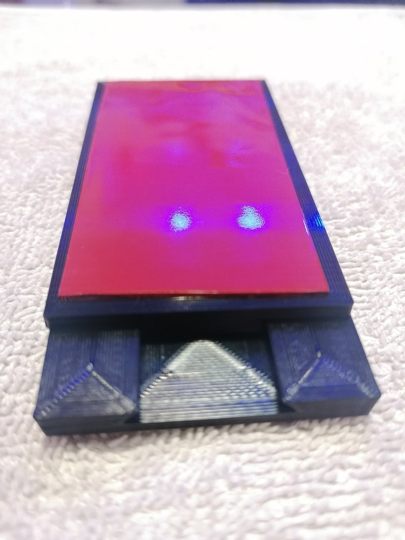 May include: A black 3D printed phone stand with a red adhesive pad on top. The stand has two triangular supports on the bottom.